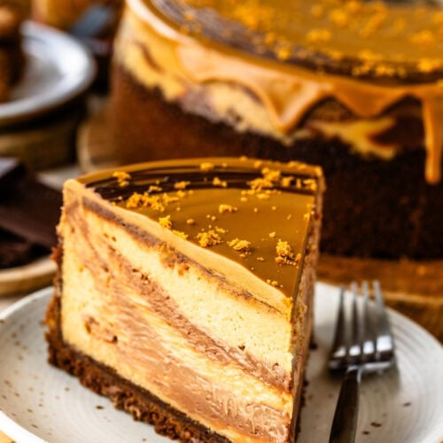 A slice of beautiful Chocolate Biscoff Cheesecake with a biscoff white chocolate ganache atop a white porcelain plate and decorated with biscoff cookie crumbs.