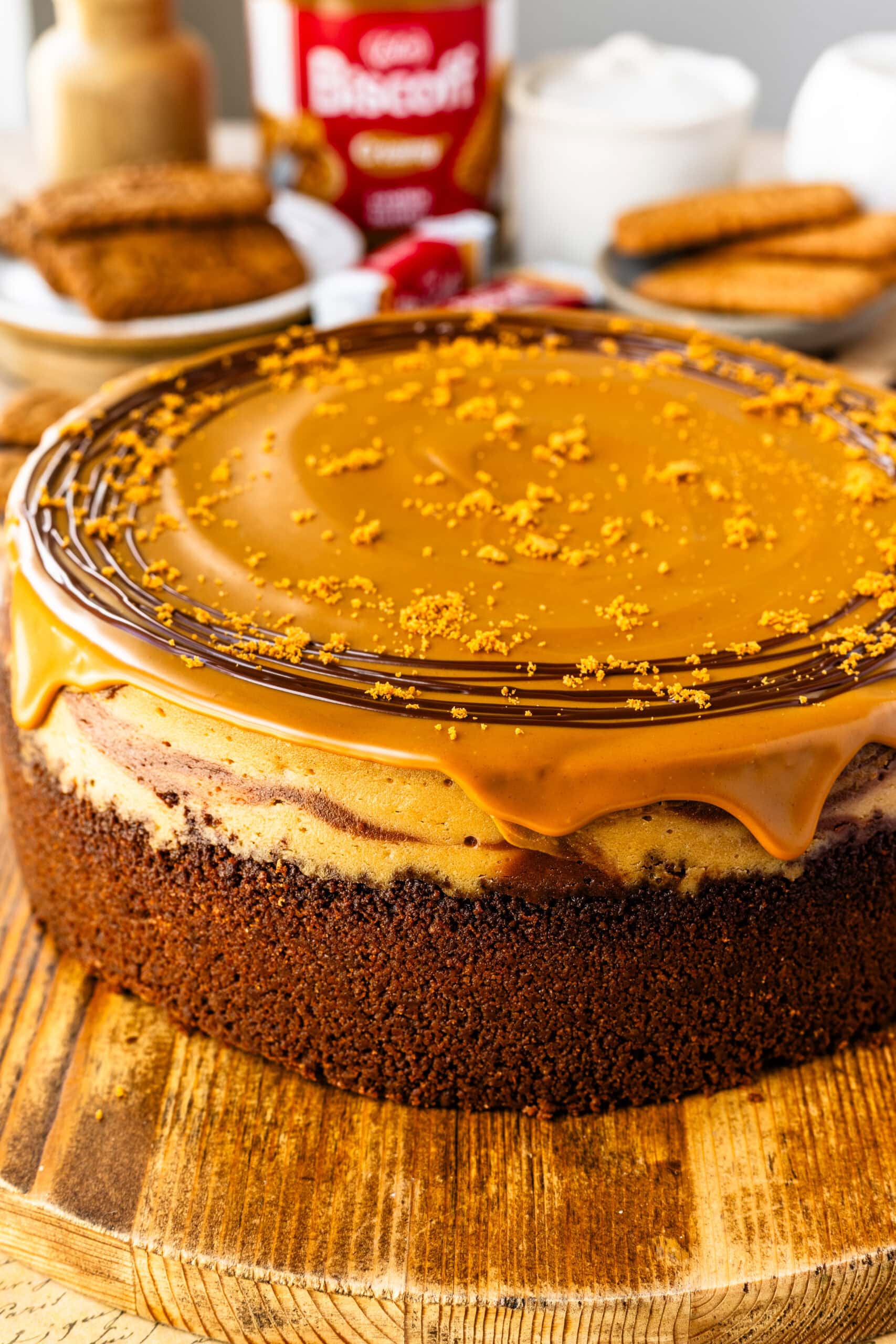 An entire, beautiful Chocolate Biscoff Cheesecake with a biscoff white chocolate ganache atop a wooden serving board and decorated with biscoff cookie crumbs.