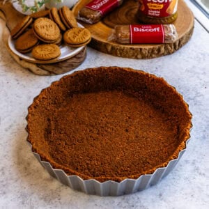 A pressed Biscoff cookie crust in a tart pan, ready for filling, with Biscoff cookies and cookie butter in the background.