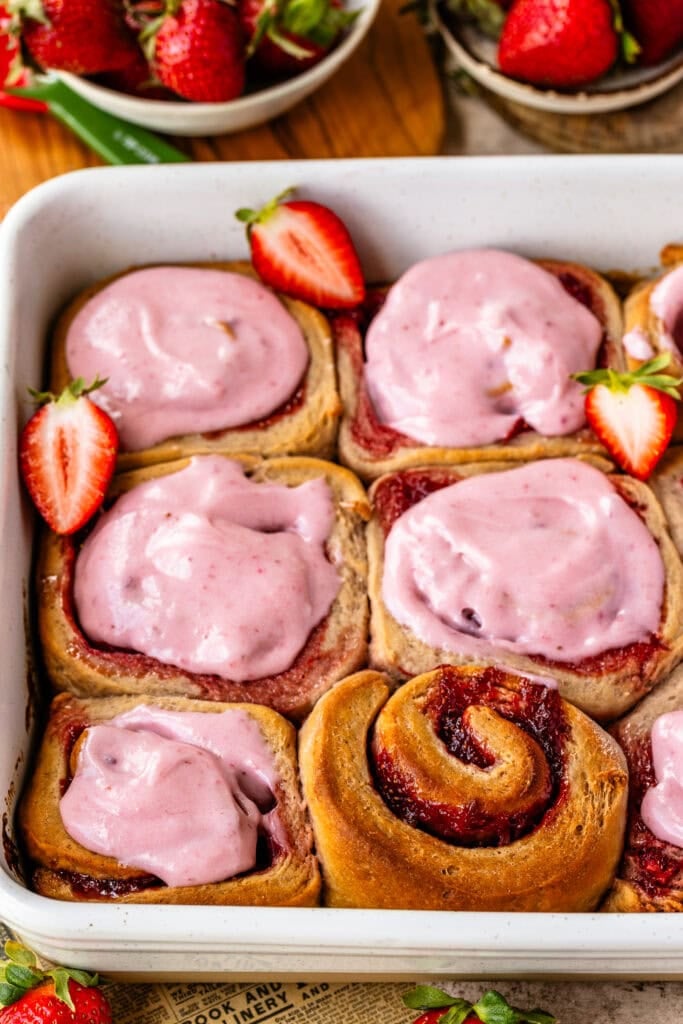 A baking dish of strawberry rolls topped with pink strawberry cream cheese frosting, with visible swirls of strawberry filling and fresh strawberries around the pan.