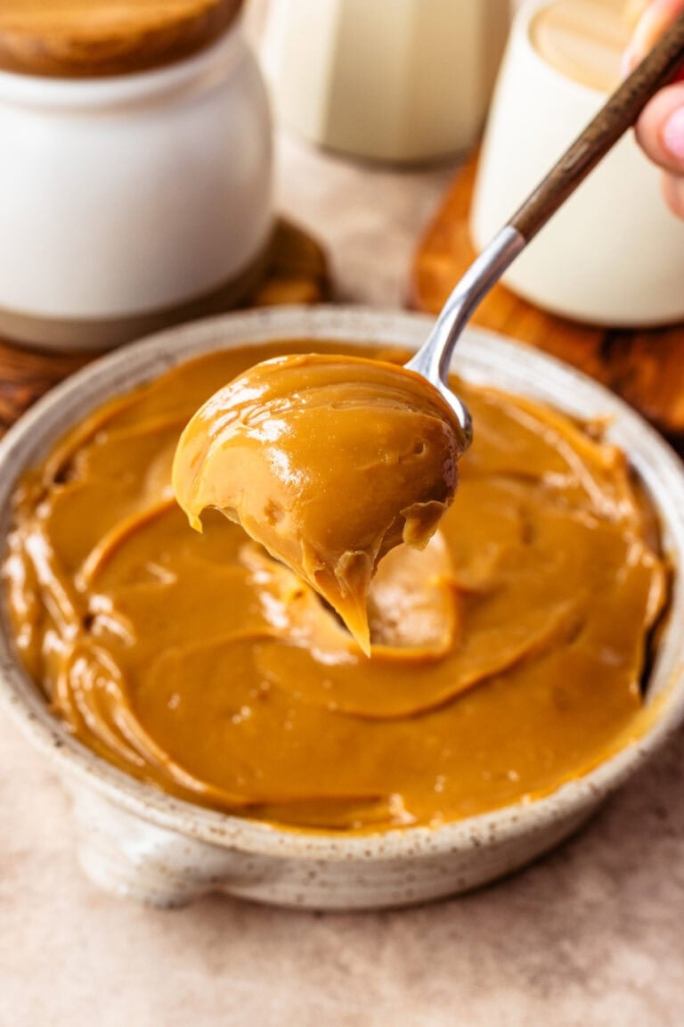 A spoon lifting thick and glossy oven-made dulce de leche from a shallow ceramic bowl.