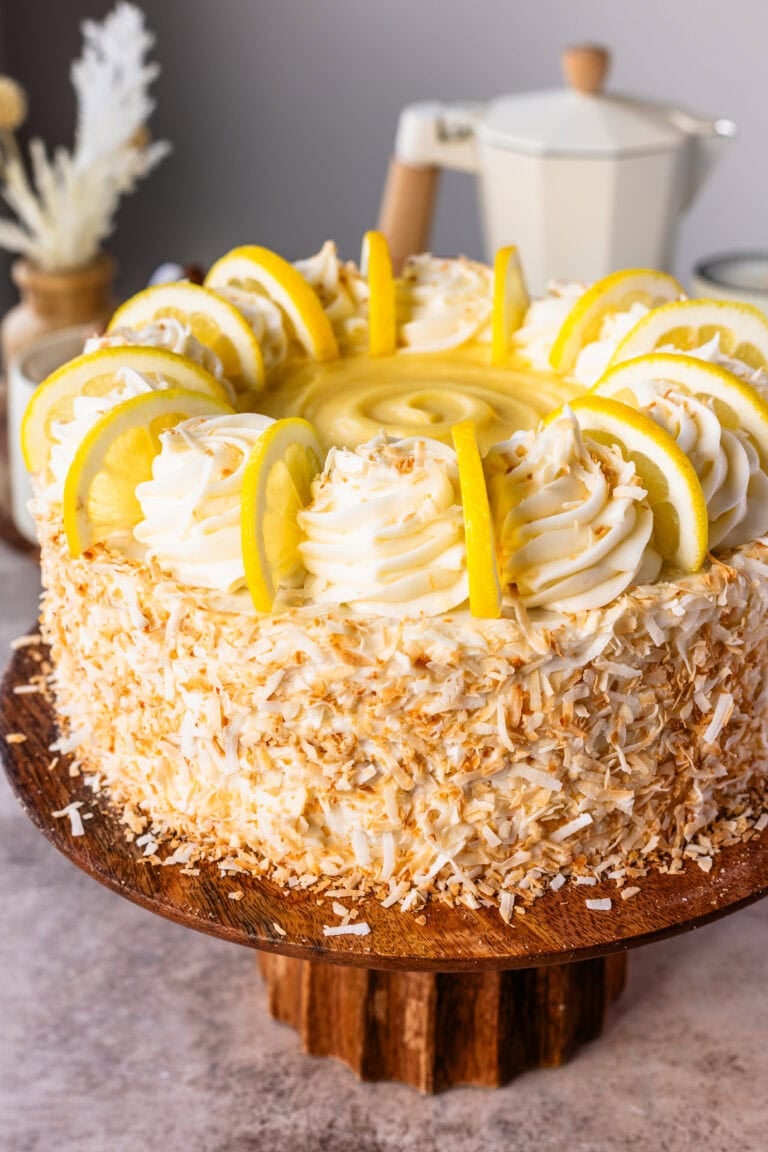 A close up of an entire Lemon Coconut Cake on a cake stand, decorated brightly with toasted coconut, lemon slices, and frosting.