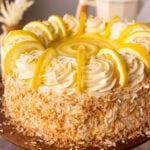 A close up of an entire Lemon Coconut Cake on a cake stand, decorated brightly with toasted coconut, lemon slices, and frosting.