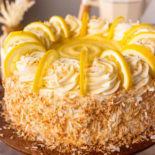 A close up of an entire Lemon Coconut Cake on a cake stand, decorated brightly with toasted coconut, lemon slices, and frosting.
