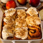 Freshly baked Peach Cobbler Cinnamon Rolls topped with cinnamon cream cheese frosting and crunchy streusel topping in a baking pan, with ripe peaches in the background.