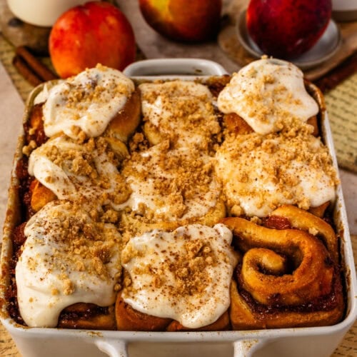 Freshly baked Peach Cobbler Cinnamon Rolls topped with cinnamon cream cheese frosting and crunchy streusel topping in a baking pan, with ripe peaches in the background.