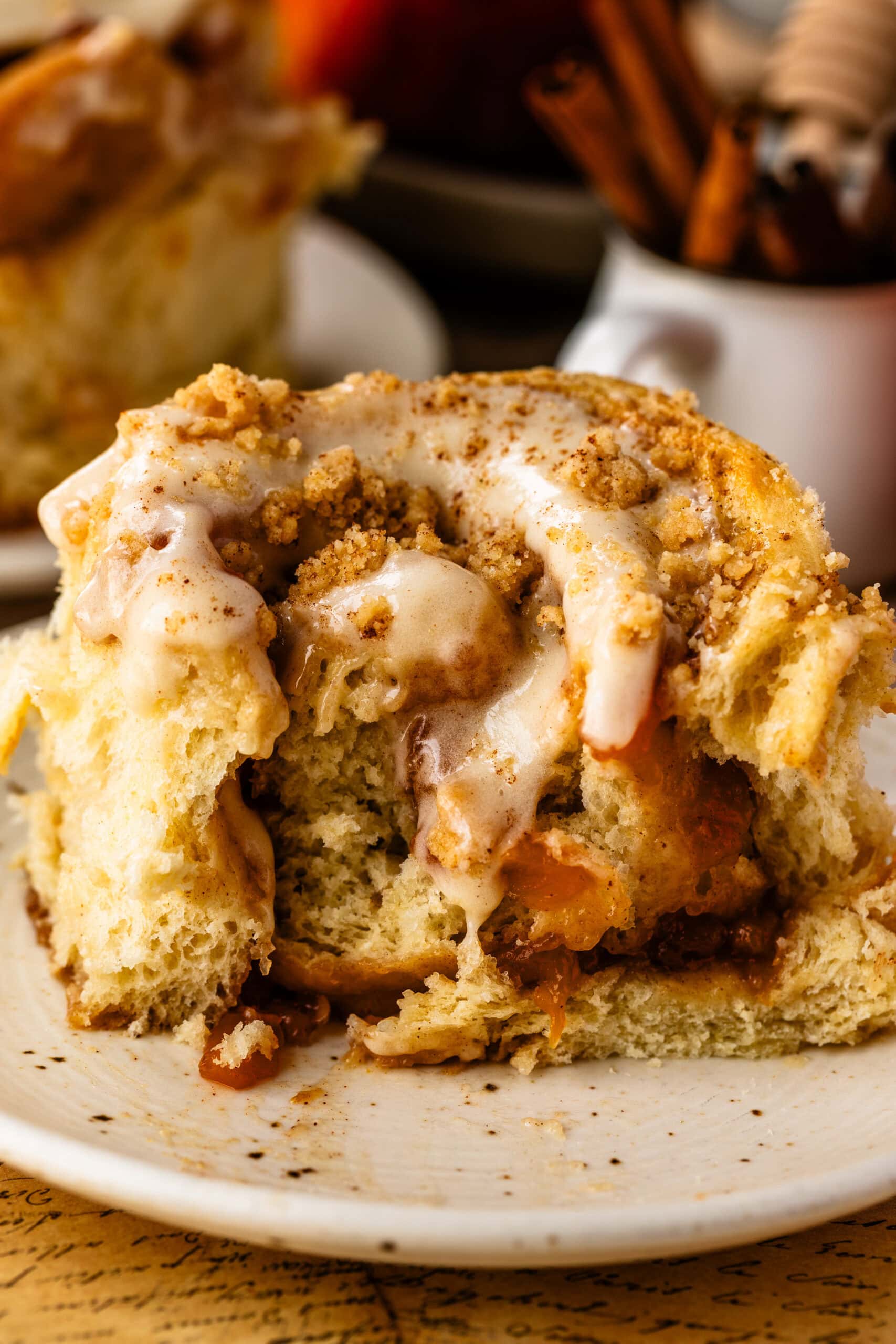 Single Peach Cobbler Cinnamon Roll on a plate with creamy cinnamon frosting and streusel topping, highlighting the tender, pillowy texture of the roll. A bite has been taken out to show that further.
