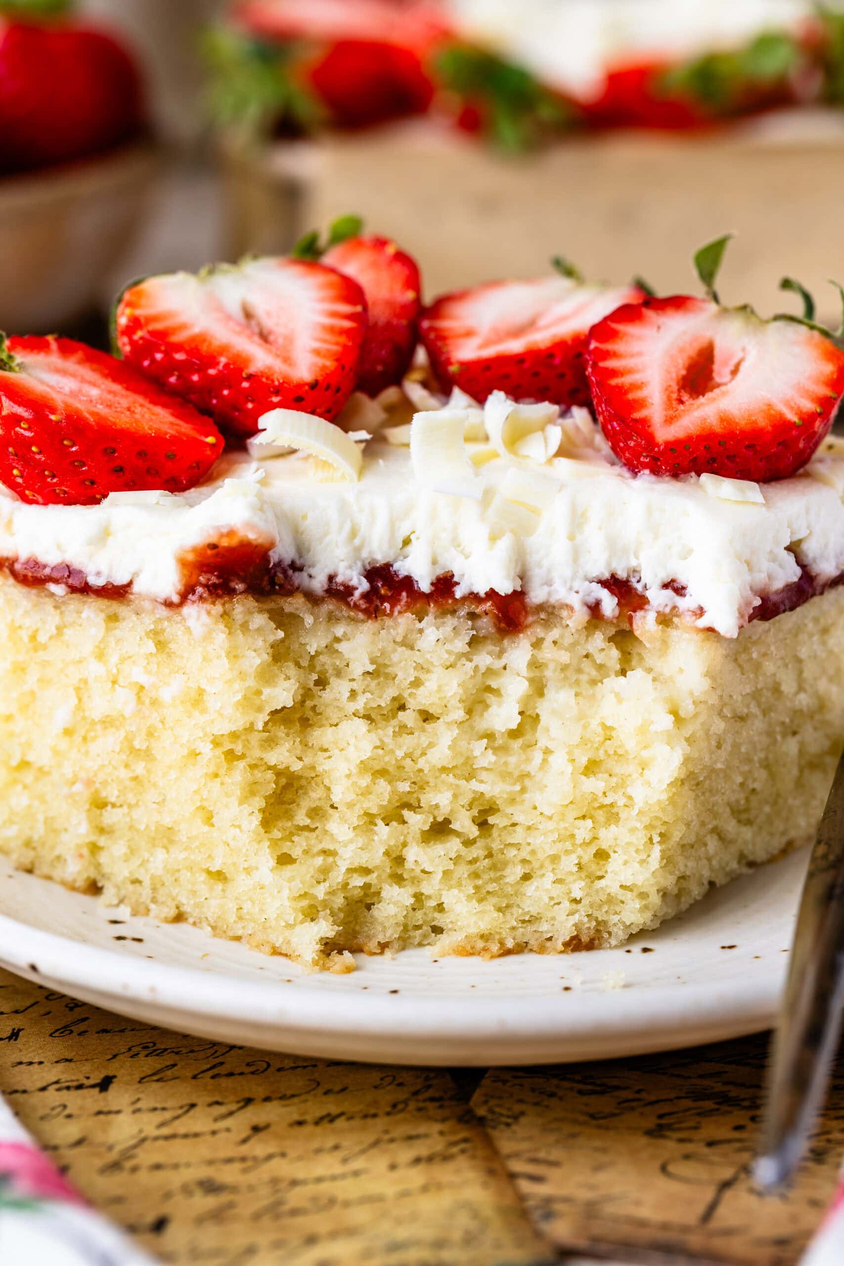 Slice of Strawberry Chantilly Sheet Cake topped with fresh strawberries, fluffy Chantilly frosting, strawberry sauce, and white chocolate curls on a dessert plate. A bite has been taken out.