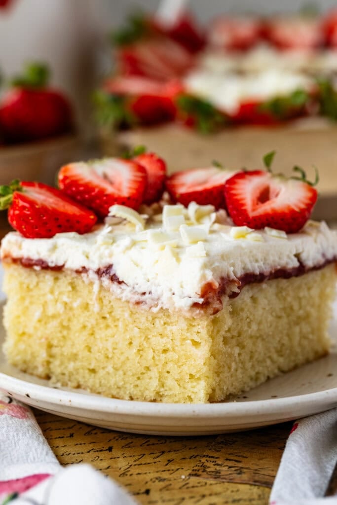 Slice of Strawberry Chantilly Sheet Cake topped with fresh strawberries, fluffy Chantilly frosting, strawberry sauce, and white chocolate curls on a dessert plate