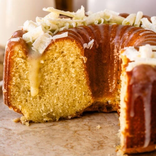 A partially sliced Vanilla Bundt Cake drizzled with white chocolate vanilla bean ganache and topped with white chocolate curls, displayed on a serving platter.