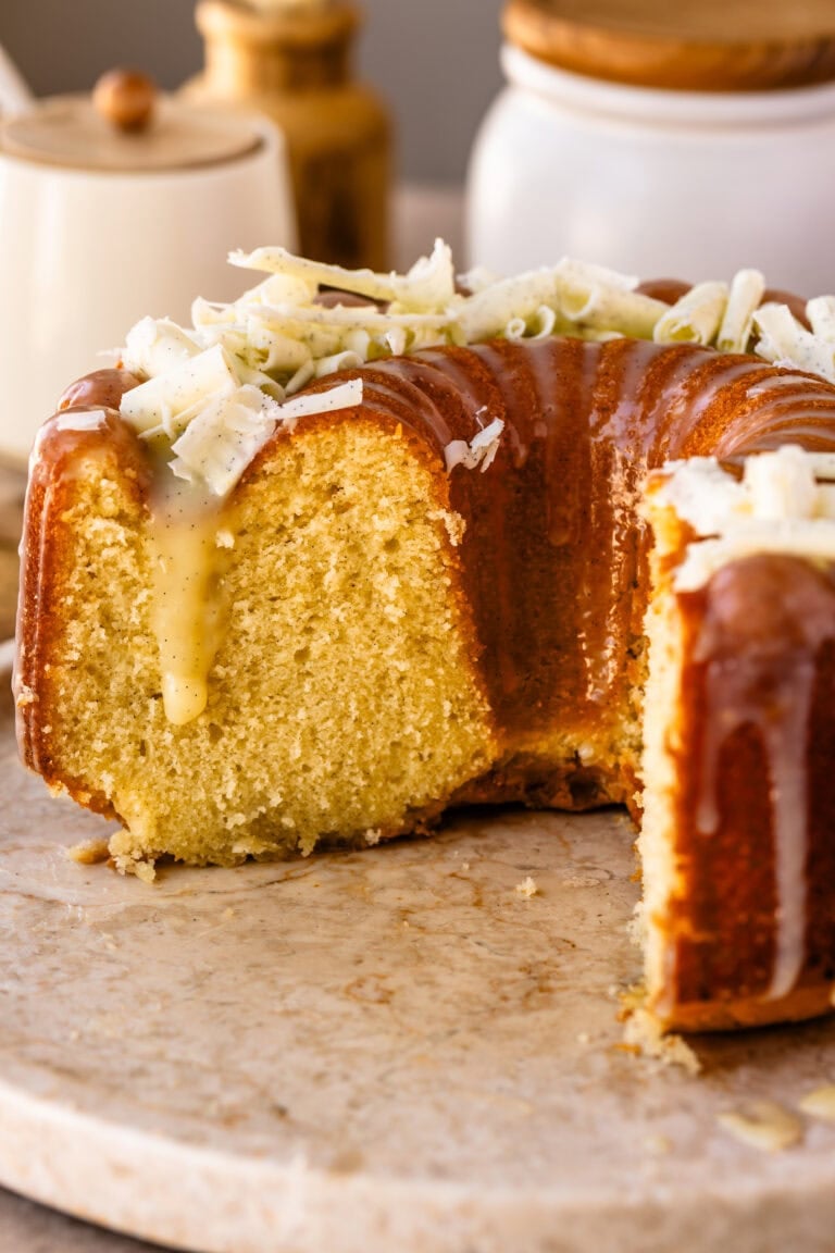 A partially sliced Vanilla Bundt Cake drizzled with white chocolate vanilla bean ganache and topped with white chocolate curls, displayed on a serving platter.