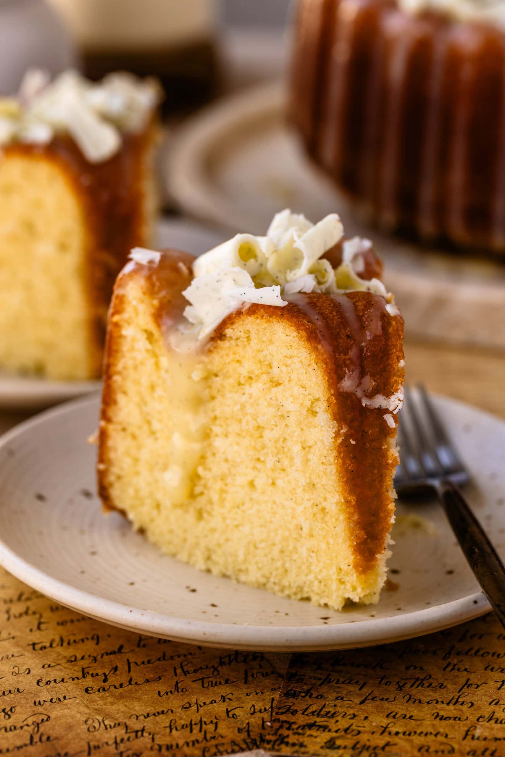 A single slice of homemade Vanilla Bundt Cake with white chocolate vanilla bean ganache dripping down the sides and garnished with white chocolate curls.