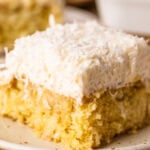A single slice of delicate coconut cake that has been soaked with a coconut milk soak, topped with coconut cloud frosting and shredded coconut.