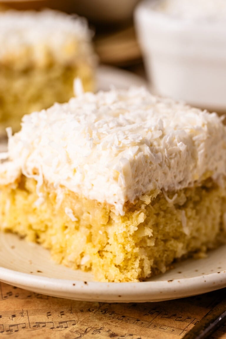 A single slice of delicate coconut cake that has been soaked with a coconut milk soak, topped with coconut cloud frosting and shredded coconut.