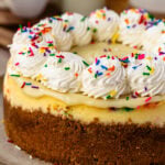 Funfetti Cheesecake decorated with whipped cream swirls, rainbow sprinkles, and white chocolate ganache.