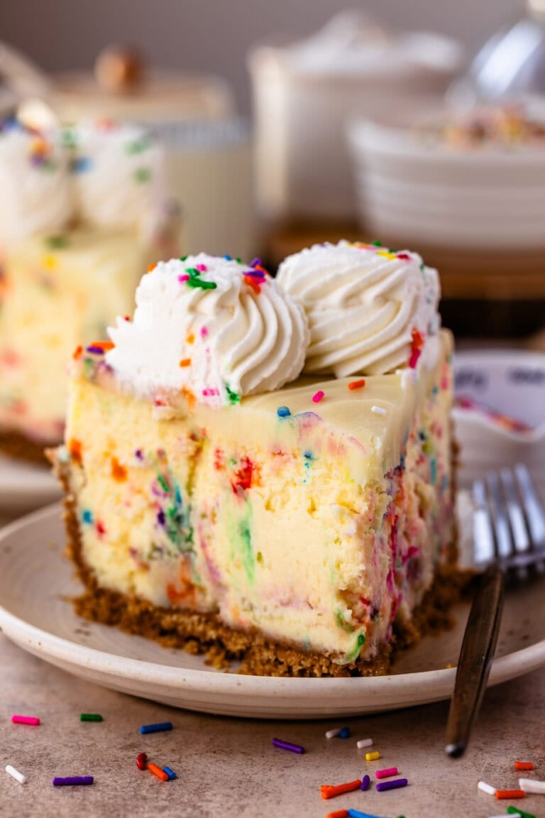 A slice of Funfetti Cheesecake with rainbow sprinkles, white chocolate ganache, and whipped cream on a graham cracker crust.