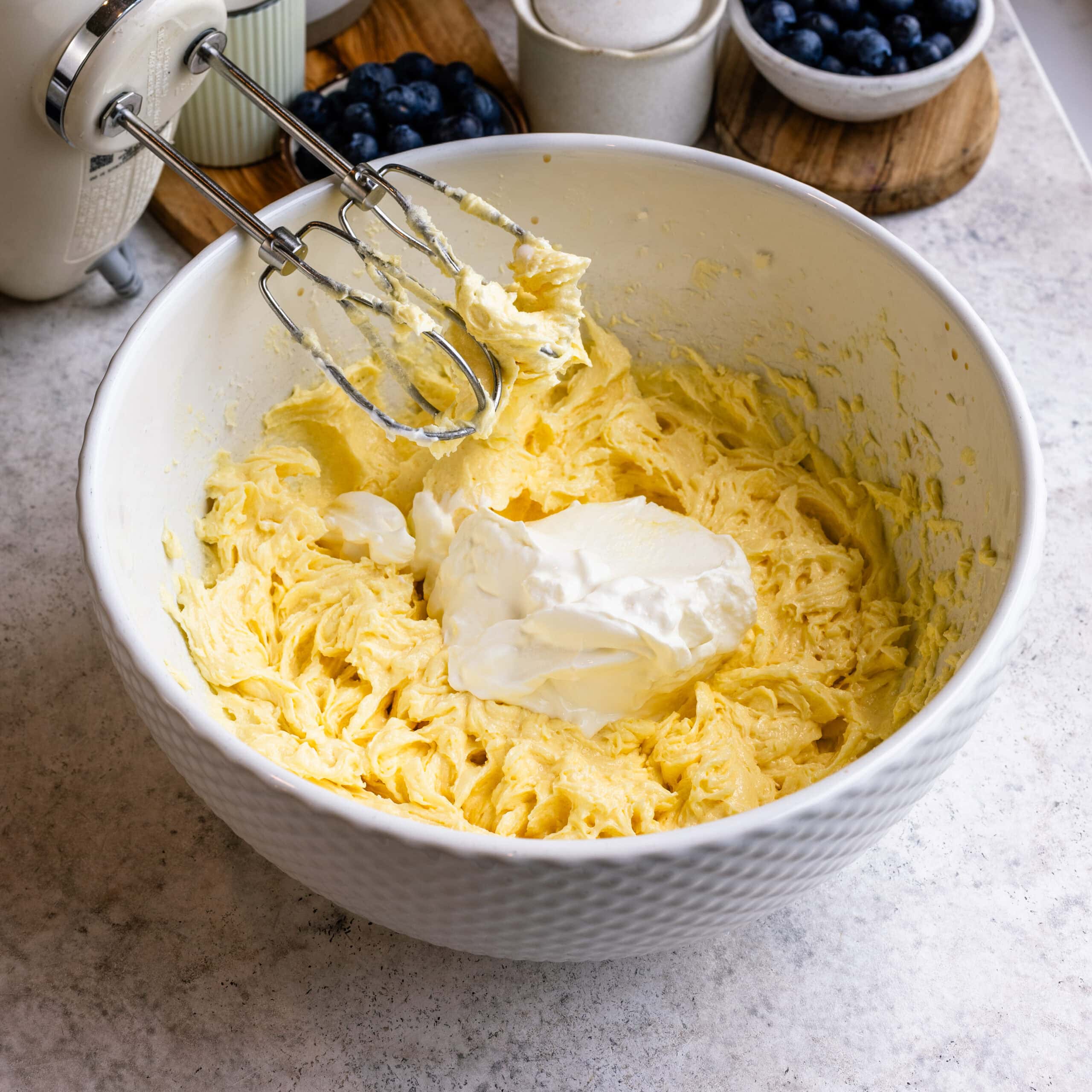 Sour cream being added to the Blueberry Sheet Cake batter in a large white mixing bowl.