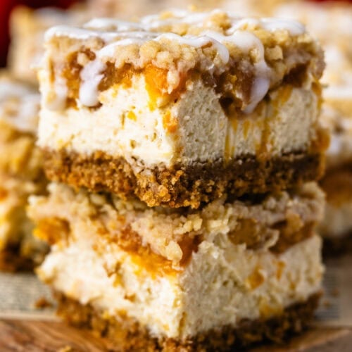 Two stacked Peach Crumble Cheesecake Bars with creamy cheesecake, spiced peach filling, buttery crumble, and cream cheese glaze.