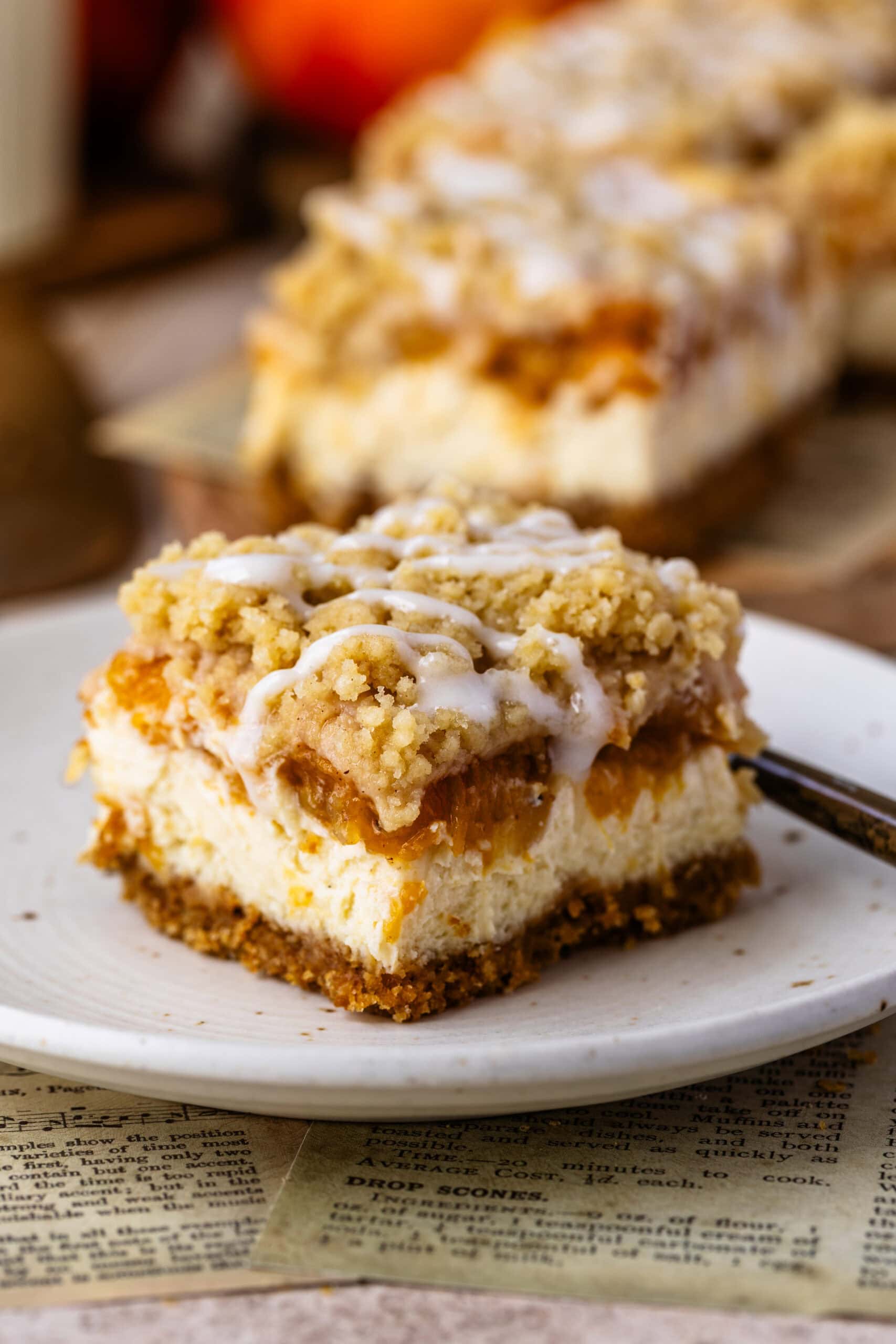 Peach Crumble Cheesecake Bar with creamy cheesecake, spiced peach filling, buttery crumble, and cream cheese glaze on a white plate.
