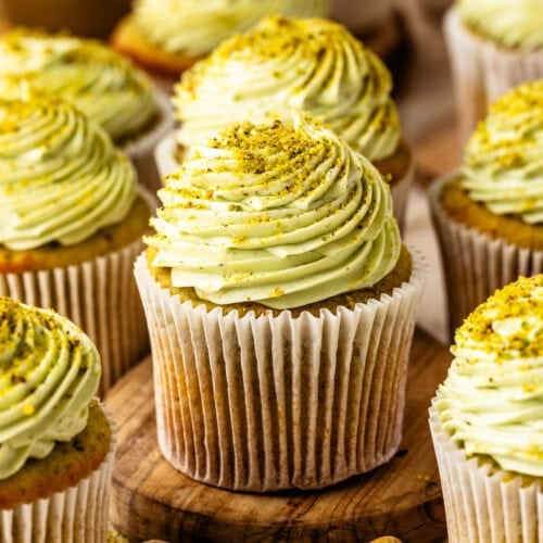 Pistachio cupcakes with pistachio cream cheese frosting and chopped pistachios on a wooden board.