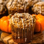 Bakery-style pumpkin muffin with a golden cinnamon crumble topping and maple glaze, surrounded by pumpkins and cinnamon sticks.