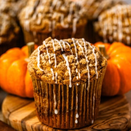 Bakery-style pumpkin muffin with a golden cinnamon crumble topping and maple glaze, surrounded by pumpkins and cinnamon sticks.